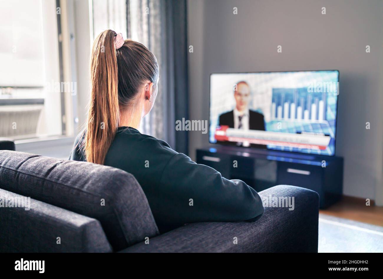 TV-Nachrichten. Frau, die im Wohnzimmer Fernsehen schaut. Reporter, Anchor und Studio-Moderator auf dem Bildschirm mit Nachrichtenüberschriften. Sitzende Person. Stockfoto