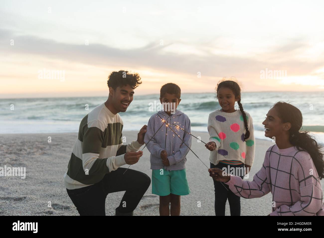 Fröhliche, multirassische Familie, die sich bei Sonnenuntergang am Strand gegen den Himmel mit Wunderkerzen vergnügt Stockfoto