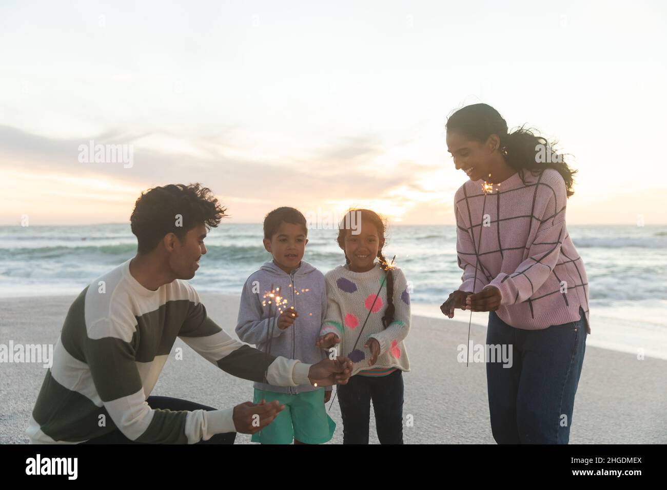Fröhliche multirassische Familie, die bei Sonnenuntergang mit Wunderkerzen am Strand gegen den Himmel genießt Stockfoto