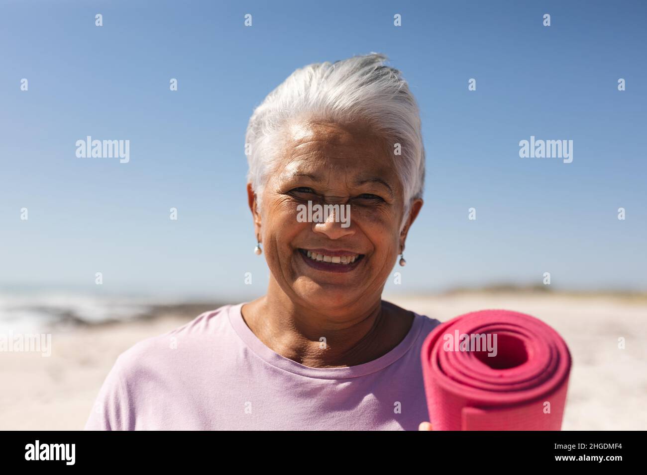 Porträt einer lächelnden älteren Frau im Ruhestand, die am Strand eine Übungsmatte mit kurzen Haaren hält Stockfoto