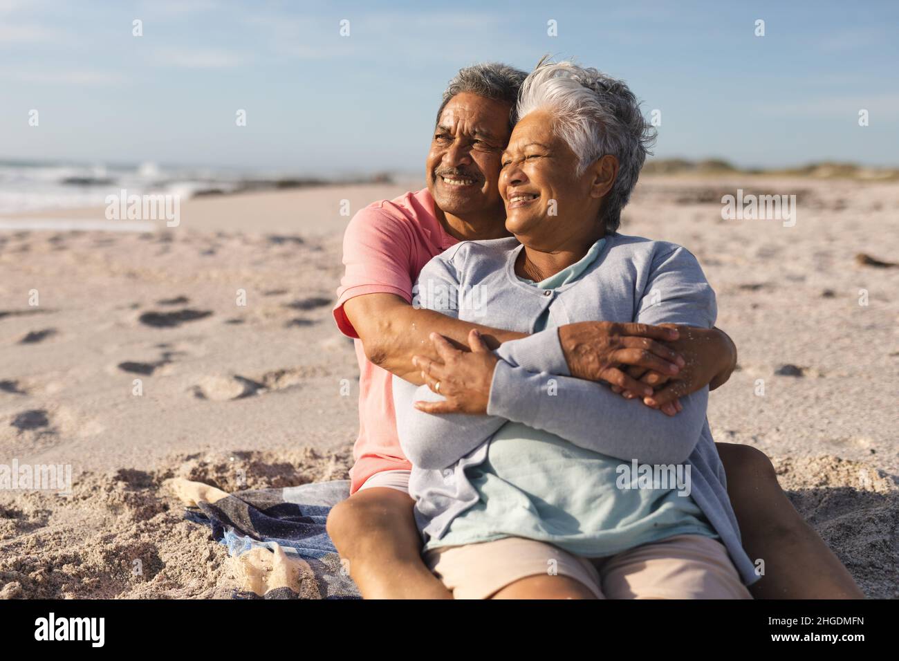 Ein lächelndes, mehrrassiges Ehepaar sitzt zusammen, während es an einem sonnigen Tag auf den Strand blickt Stockfoto