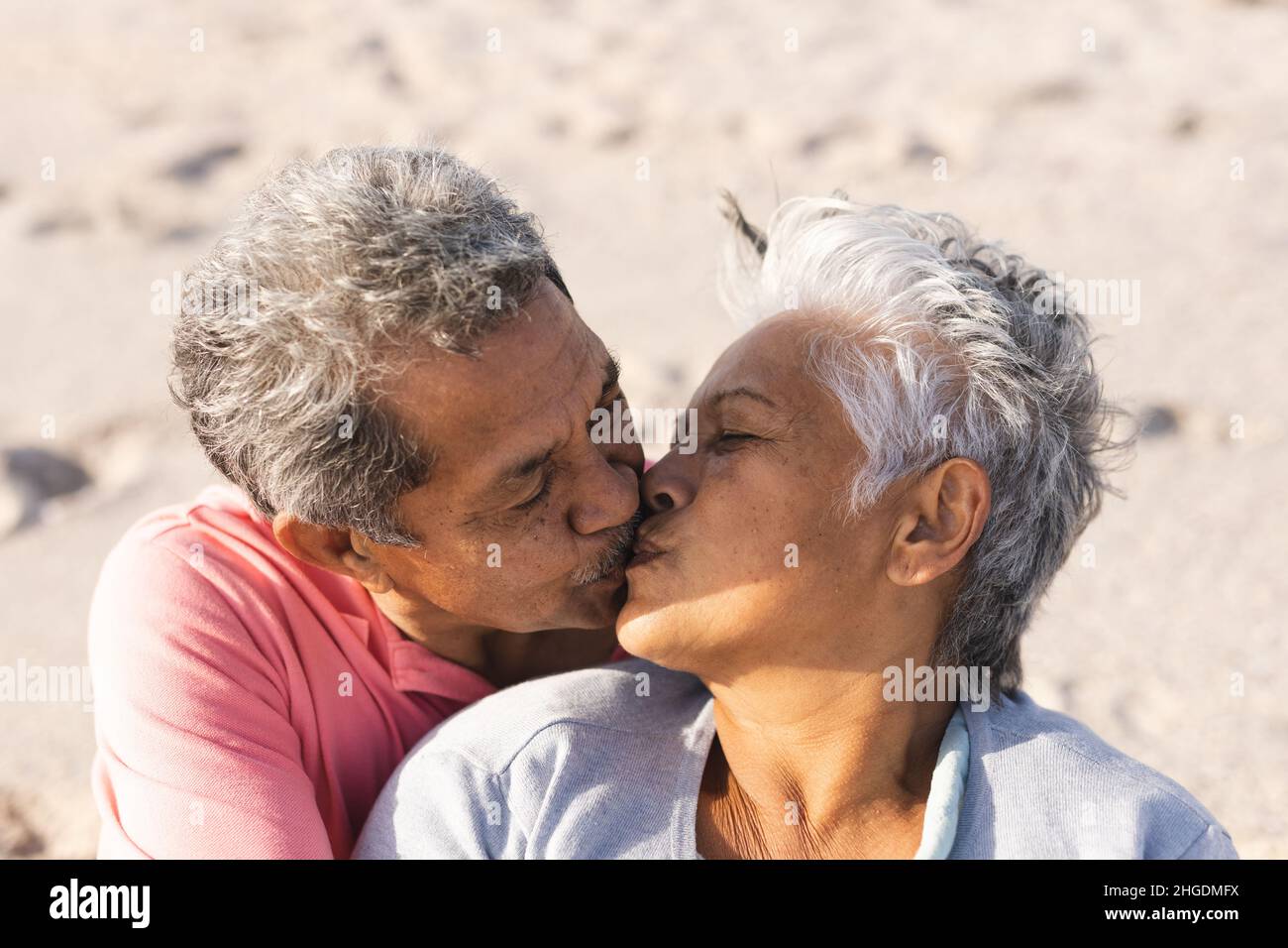 Liebevolles multirassisches Seniorenpaar, das sich auf die Lippen küsst, während es einen sonnigen Tag am Strand genießt Stockfoto