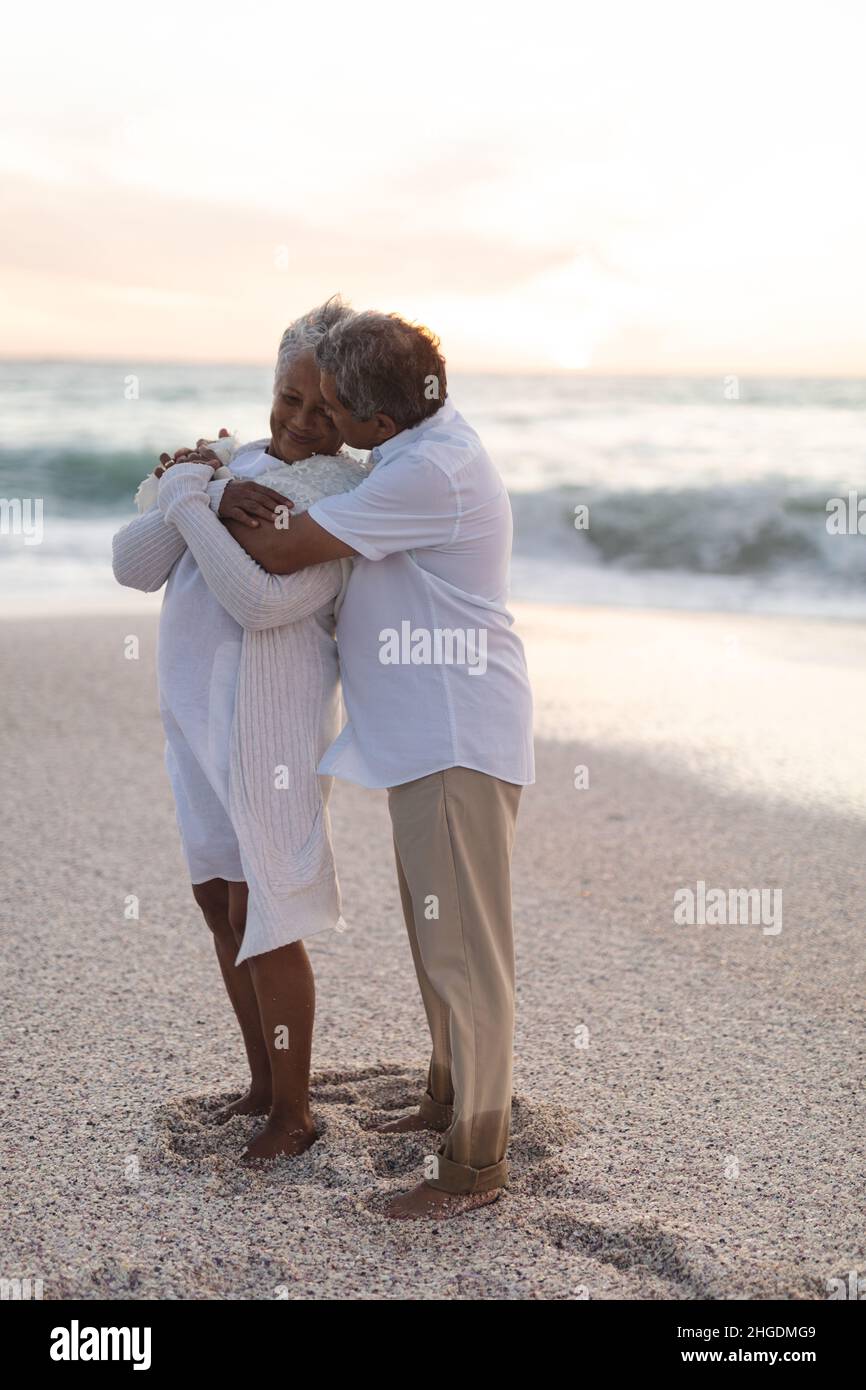 Die ganze Länge der liebevollen Senior biracial Mann umarmt Frau von hinten am Strand während des Sonnenuntergangs Stockfoto