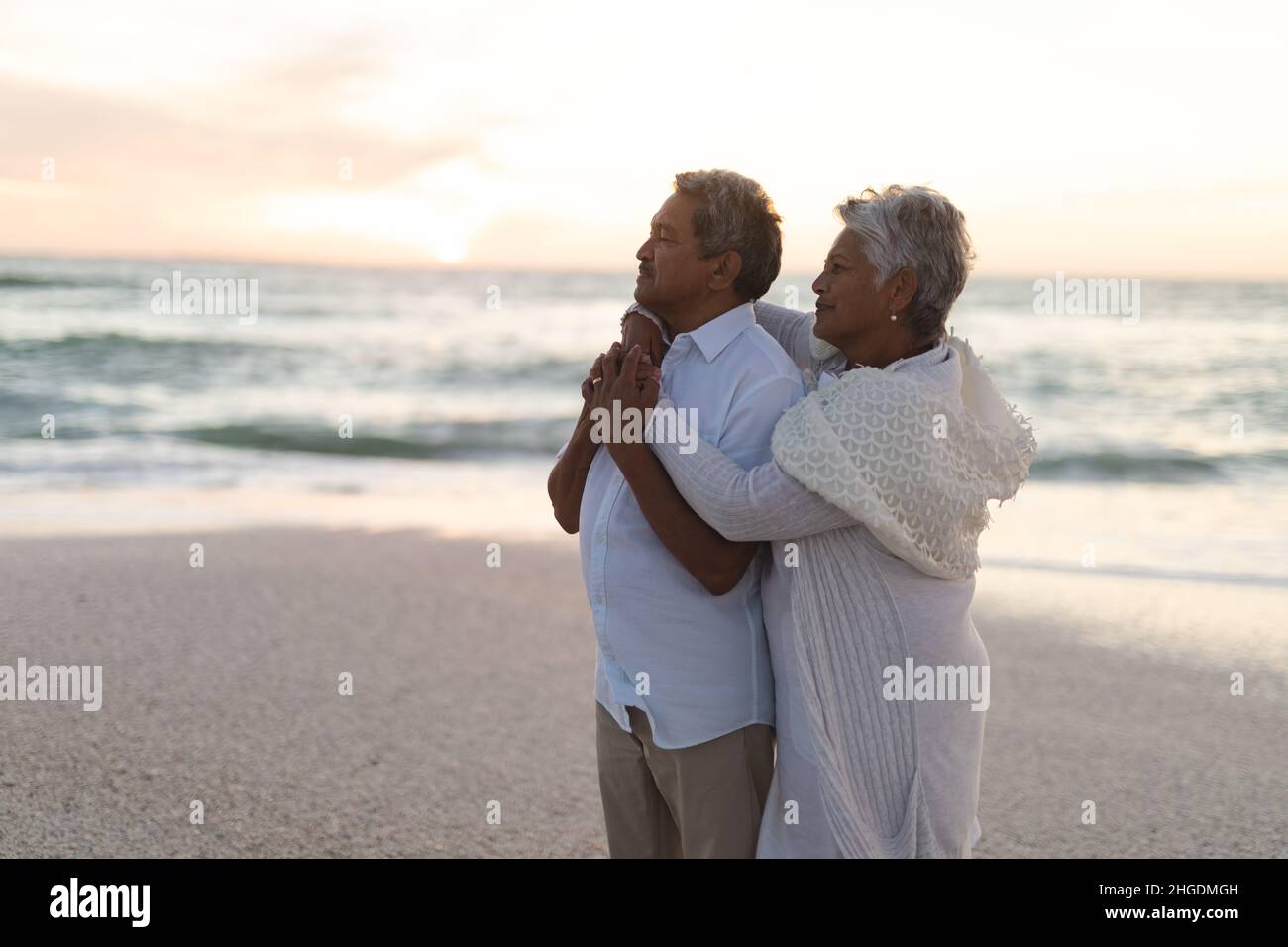 Seitenansicht eines mehrrassigen älteren Ehepaares, das während des Sonnenuntergangs am Strand umarmt und wegschaut Stockfoto