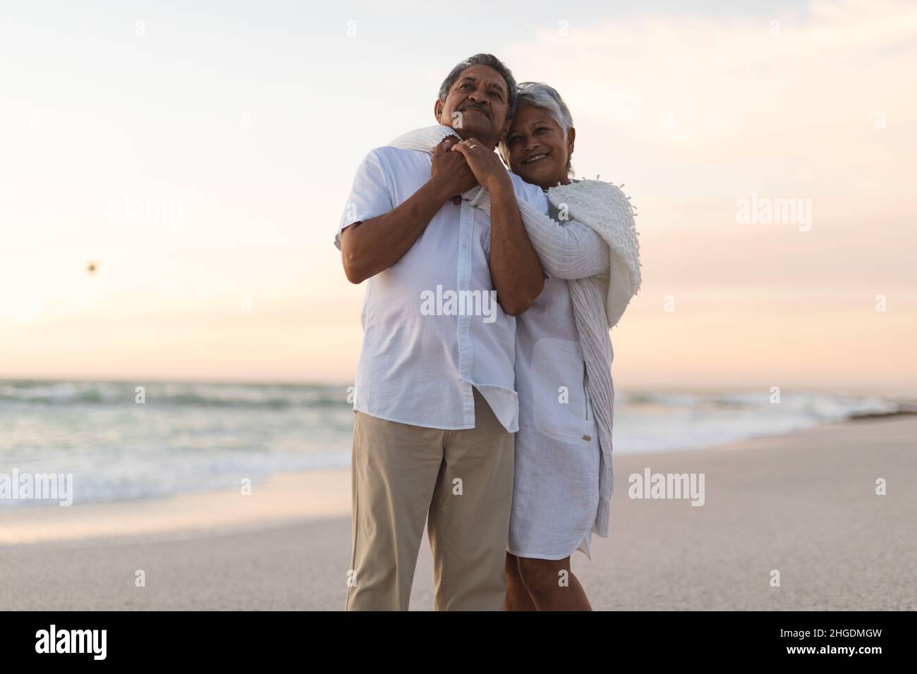 Ein glückliches, frisch verheiratetes, mehrrassiges Paar, das sich während des Sonnenuntergangs am Strand gegen den Himmel umarmt Stockfoto