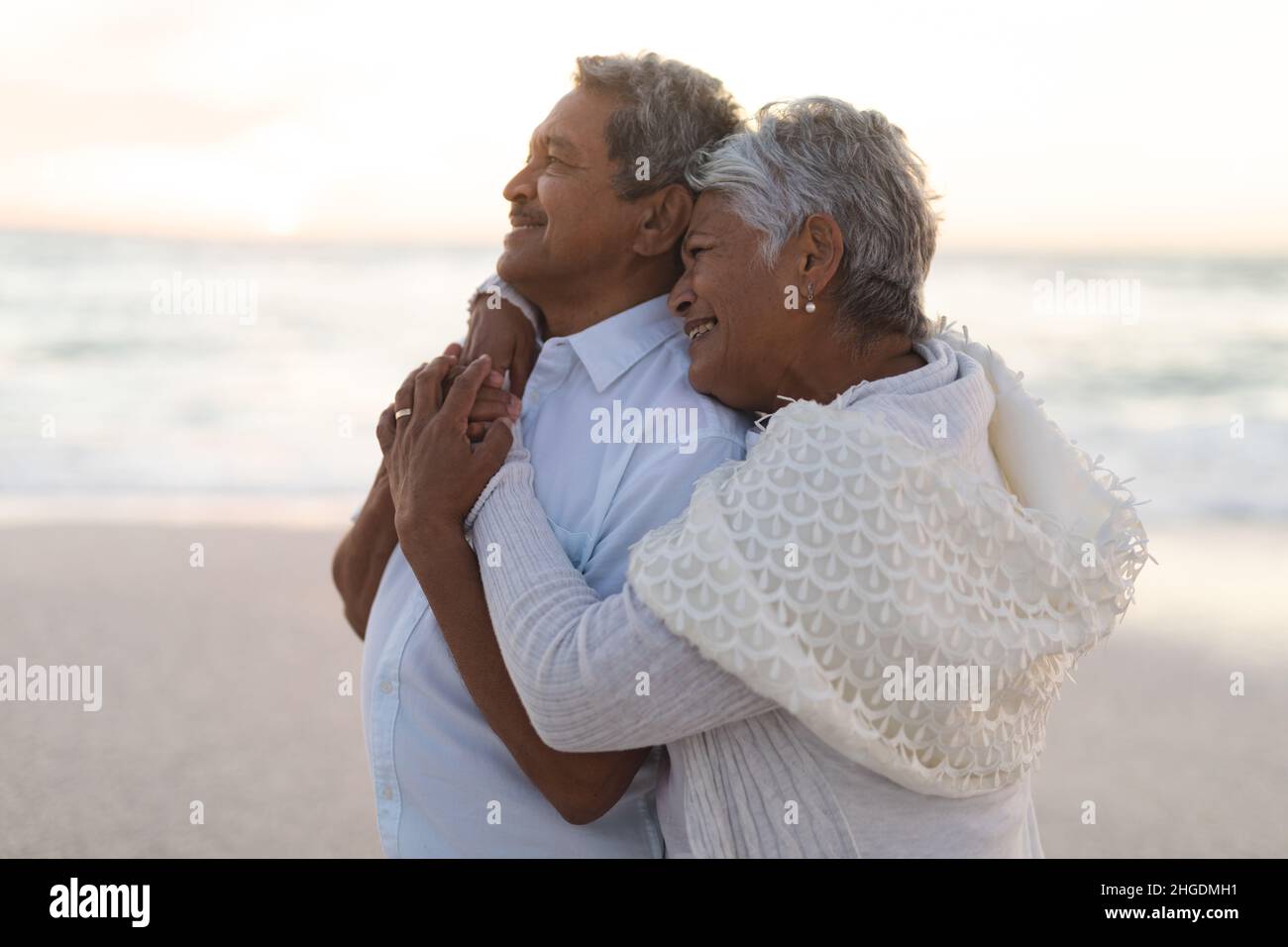 Lächelndes, frisch verheiratetes, mehrrassiges Ehepaar schaute weg, während es sich während des Sonnenuntergangs am Strand umarmte Stockfoto