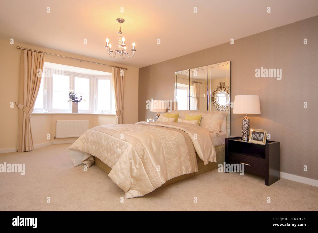 Hauptschlafzimmer in cremefarbenen neutralen Farben, beigefarbener Teppich, Kingsize-Doppelbett, Bettüberwurf, Spiegelkopfteil, Nachttischlampen, Erkerfenster. Stockfoto