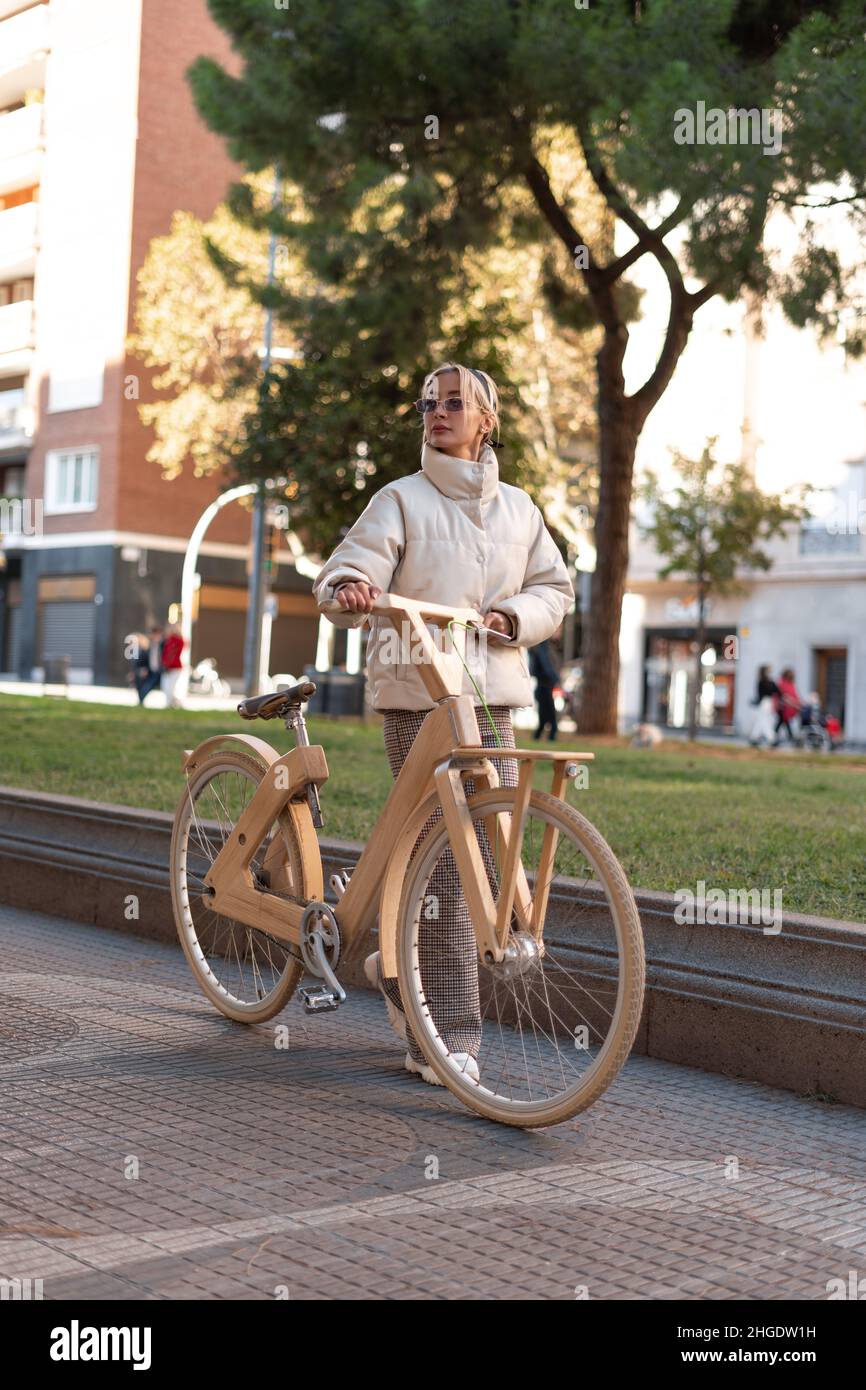 Ganzkörperweibchen in Oberbekleidung schieben Holz Öko-Fahrrad und wegschauen, während zu Fuß auf gepflasterten Weg an sonnigen Tag im Park. Stockfoto