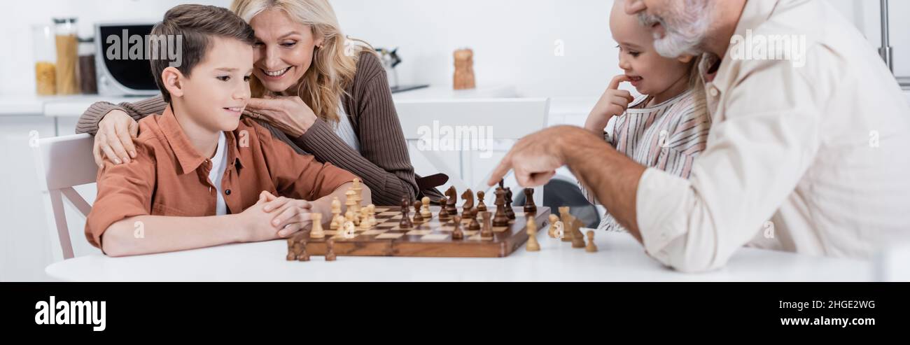 Mann zeigt auf Schachbrett, während er mit Enkeln und Frau Schach spielt, Banner Stockfoto