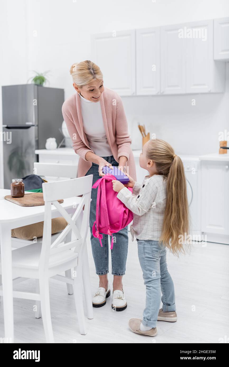 Lächelnde Frau hält die Lunchbox, während sie den Rucksack der Enkelin in der Küche packt Stockfoto