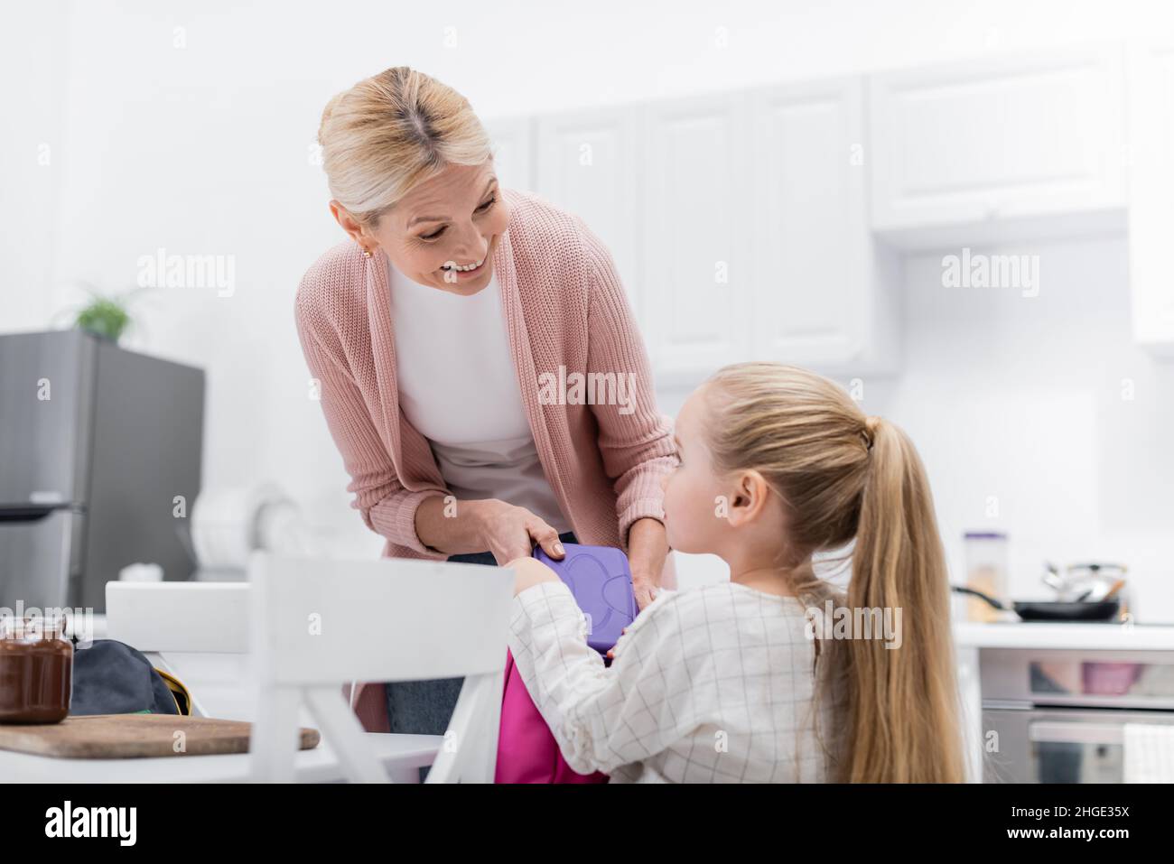 Fröhliche Frau hält die Lunchbox in der Nähe der Enkelin und ein Glas mit Schokoladenpaste in der Küche Stockfoto