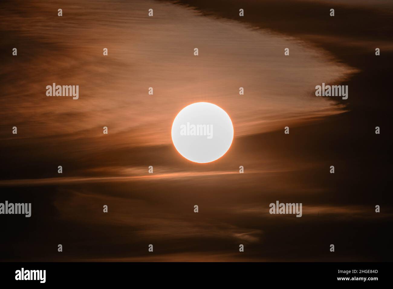Leicht bewölkt, rot getönter Himmel. In der Mitte sieht man eine helle Sonnenscheibe. Stockfoto