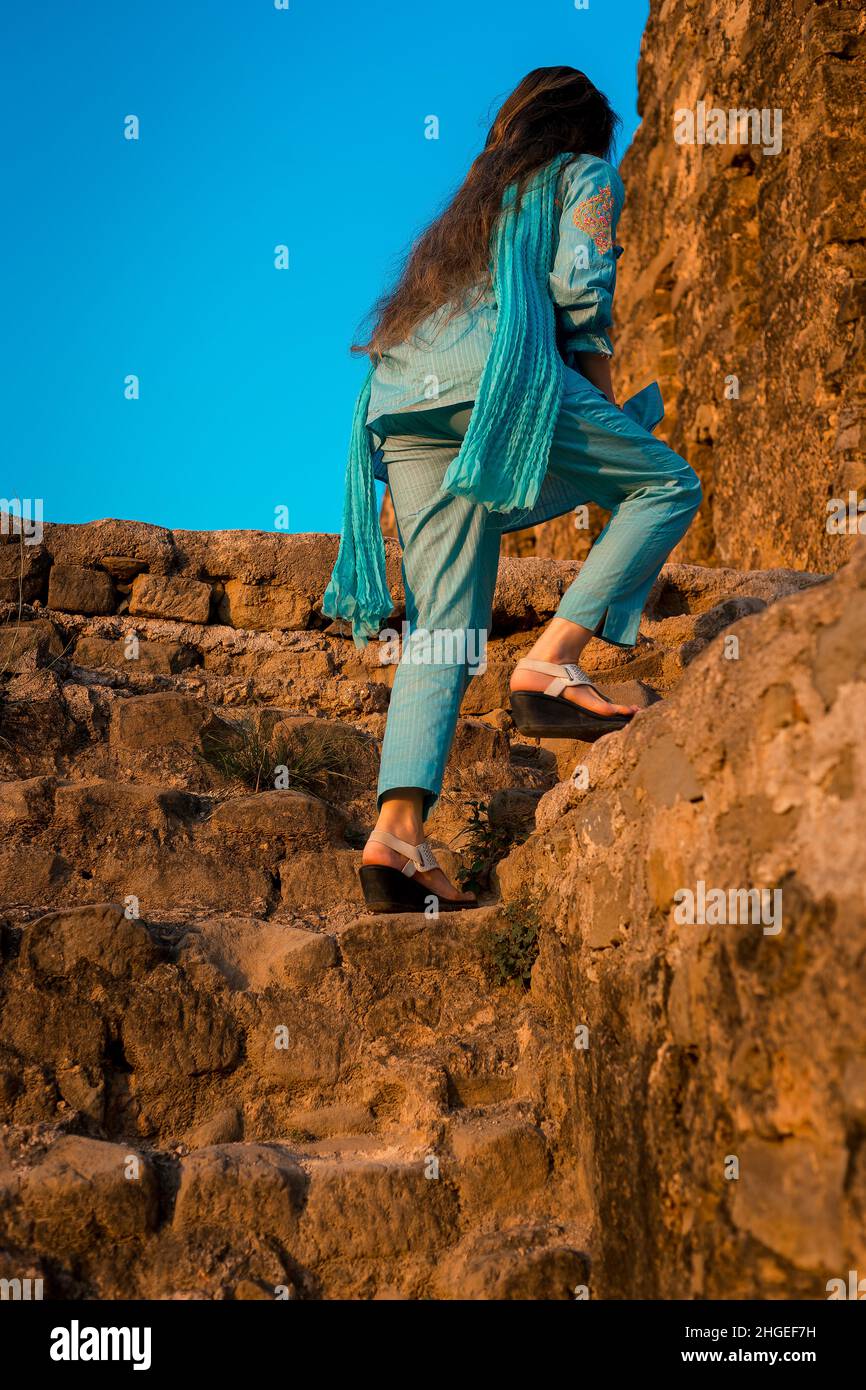 Mädchen in blauem pakistanischem Kleid mit welligem langen Haar, das tagsüber die alten Treppen des alten Fort Rohtas mit hellblauem Himmel bestiegen hat. Stockfoto