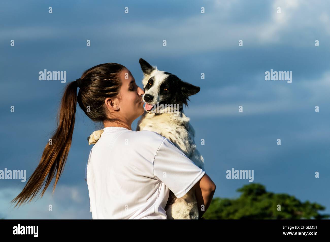 Das lateinische Mädchen hält ihren schwarz-weißen Hundefreund in den Armen und küsst sie. Das Mädchen hat einen Pony-Schwanz. Stockfoto