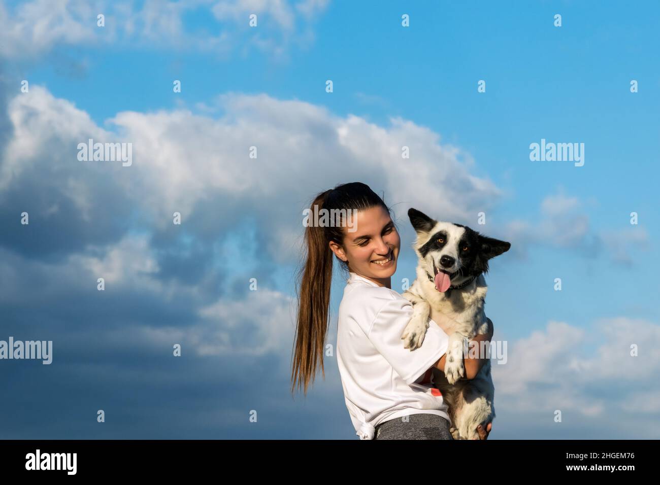 Die junge lateinische Frau hält ihren Hundefreund in den Armen und lächelt sie an. Der Hund blickt auf die Kamera, Wolken und blauer Himmel im Hintergrund Stockfoto