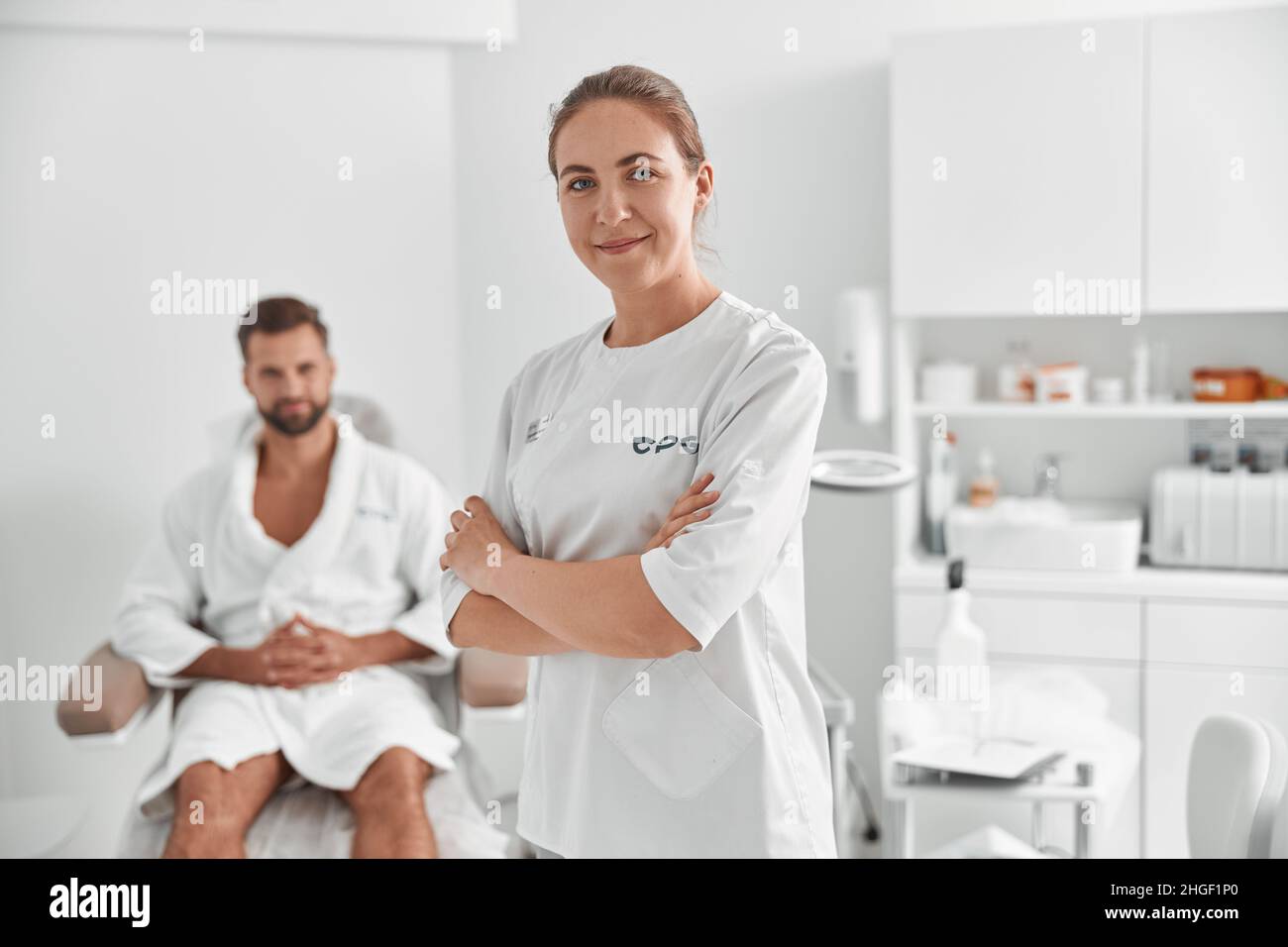 Porträt eines positiven Kosmetologen in weißer Uniform gegen verschwommene Sicht des Mannes Patienten im Schönheitssalon Stockfoto
