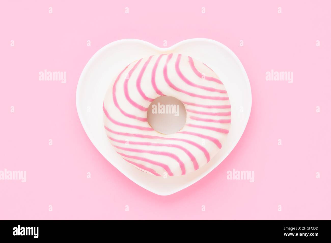 Gestreifter rosa Donut in herzförmiger Platte auf rosa Hintergrund. Valentinstag-Konzept. Draufsicht, Platz für Text kopieren. Stockfoto