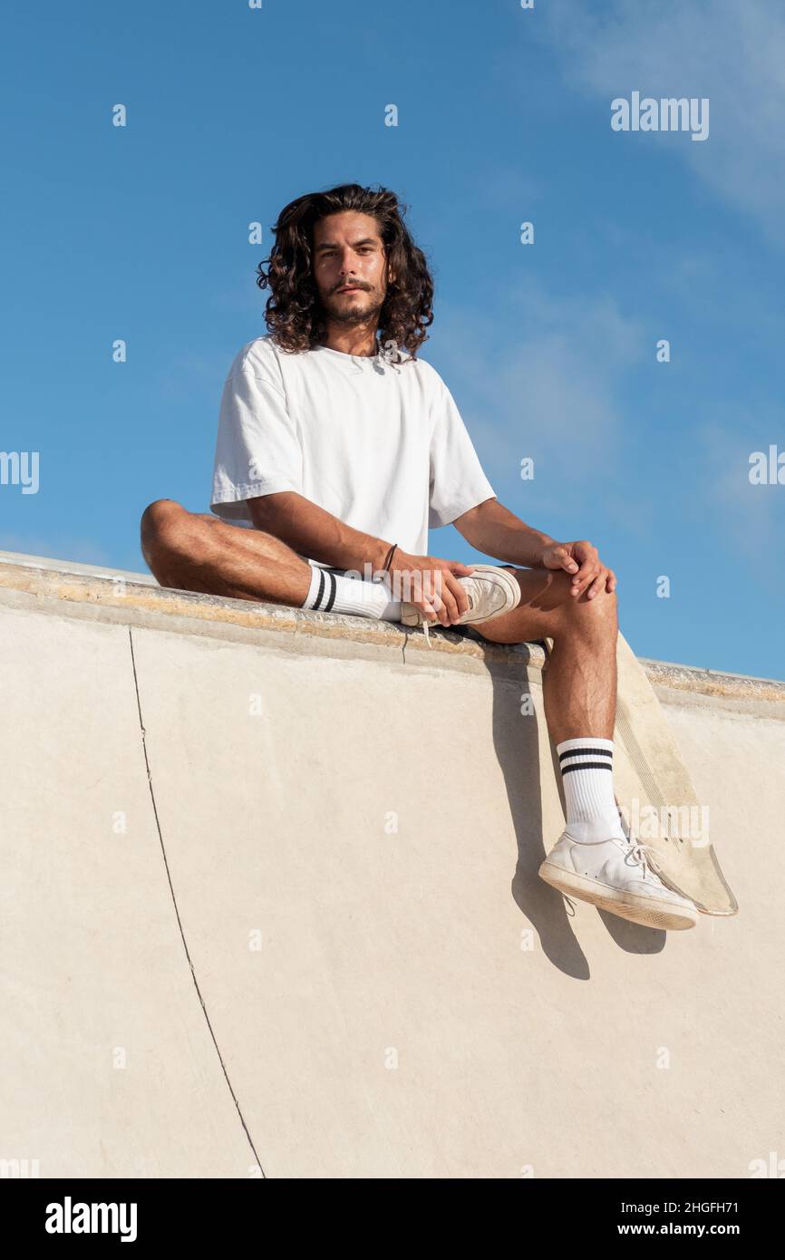 Junger Skater mit langen schwarzen Haaren sitzt am Rand der Skaterampe Stockfoto