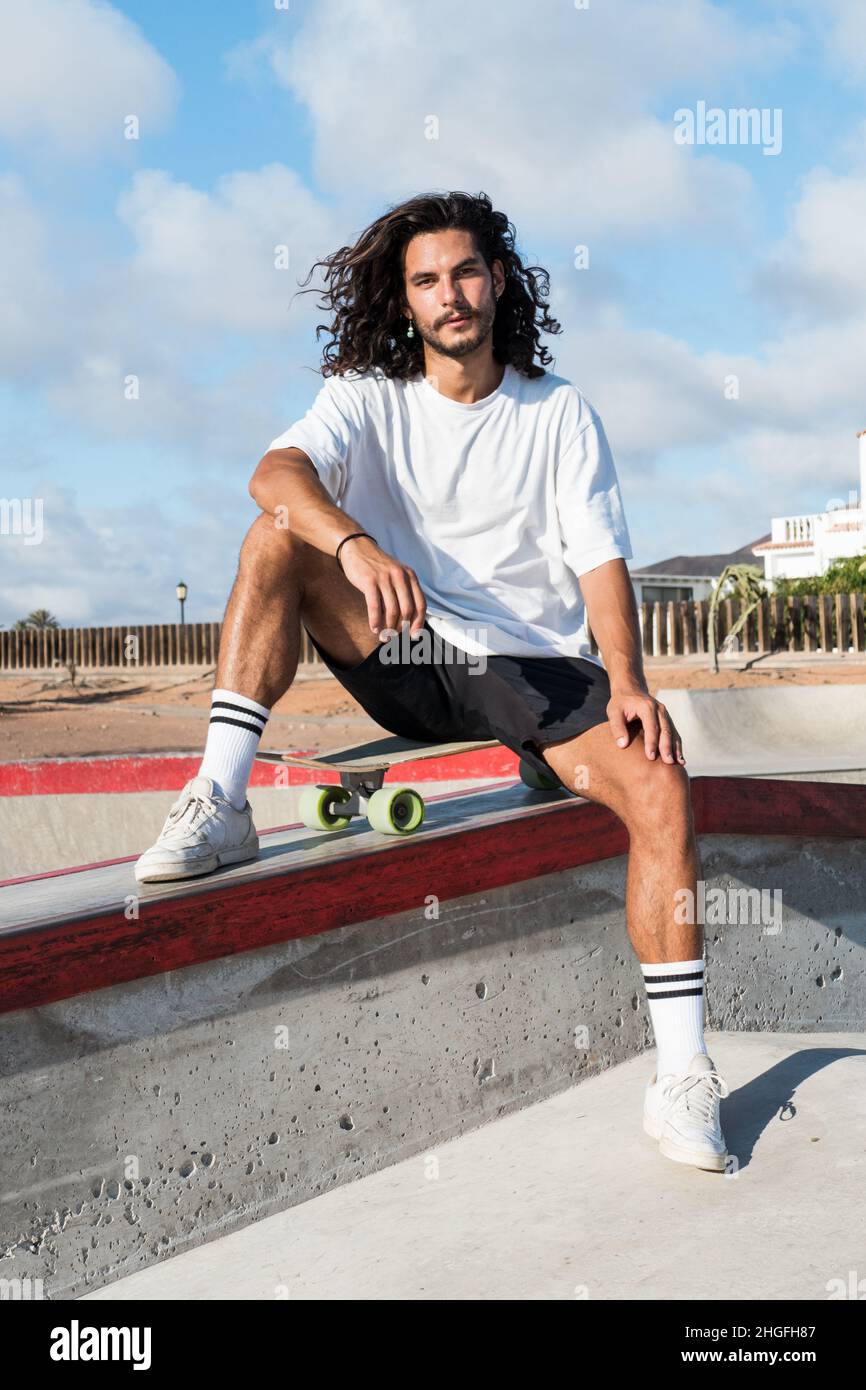 Junger, hübscher Skater, der auf seinem Skateboard im Skatepark sitzt. Er trägt Sommerkleidung und hat lange schwarze Haare und einen Bart. Stockfoto