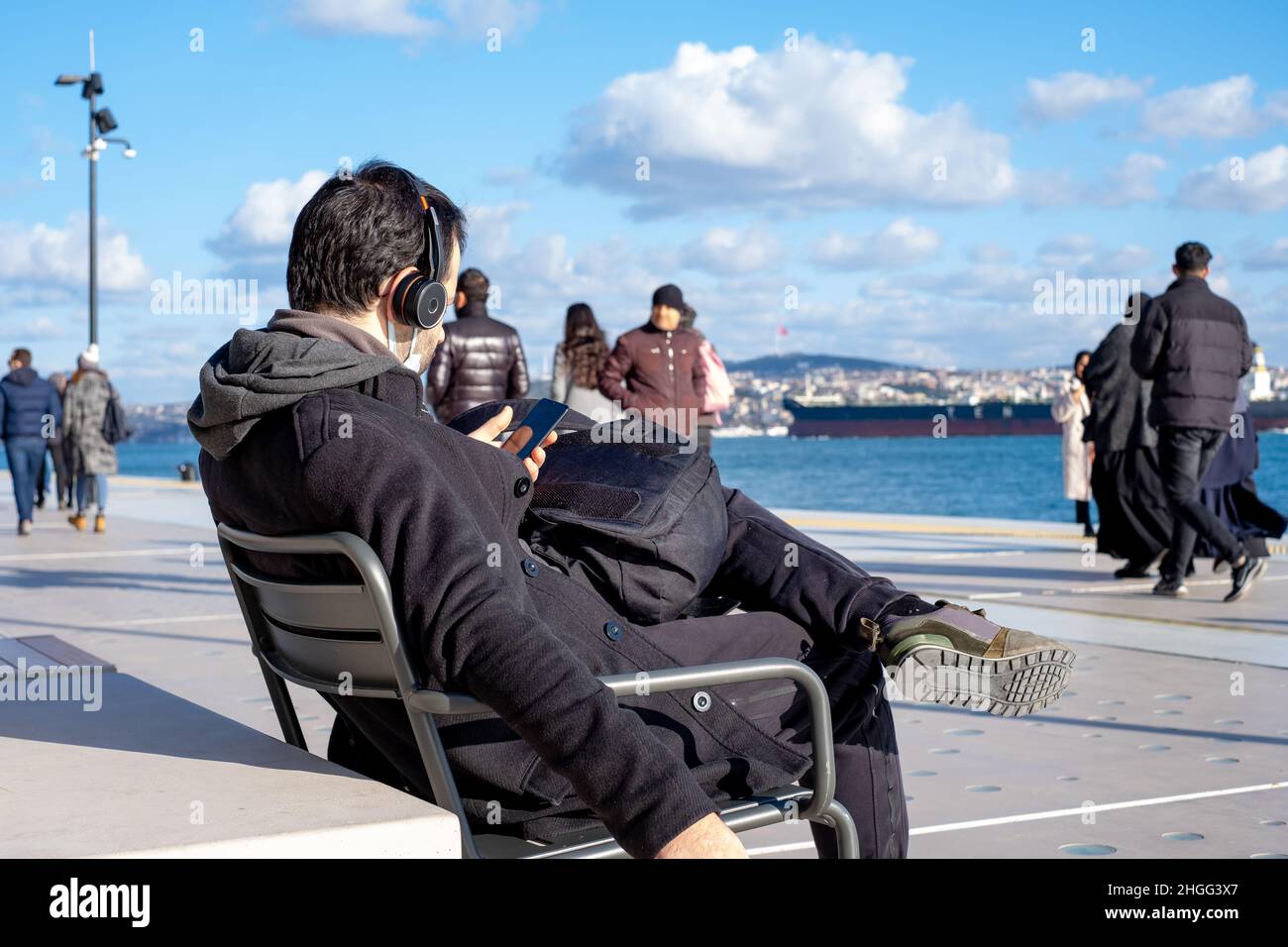 Mann, der am Meer sitzt und auf sein Telefon schaut. Mann, der am Meer sitzt und auf sein Telefon schaut, ist vor dem Hintergrund Meer und wolkenverhaufiger Himmel. Stockfoto