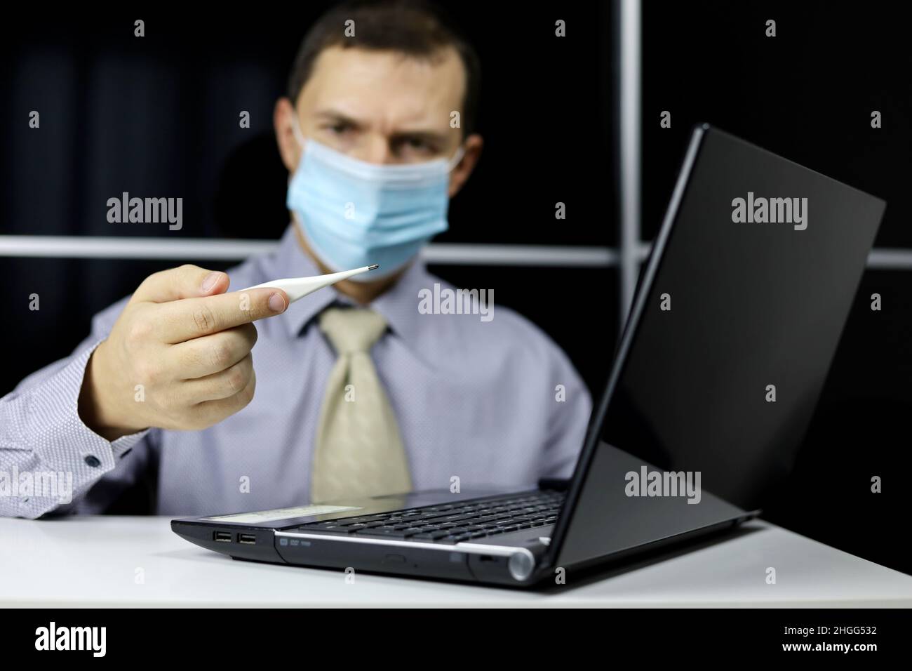Besorgter Mann in Gesichtsmaske und Bürokleidung blickt auf Digitalthermometer in der Hand, der am Laptop sitzt. Hohe Körpertemperatur, Sicherheit bei der Arbeit bei Covid Stockfoto