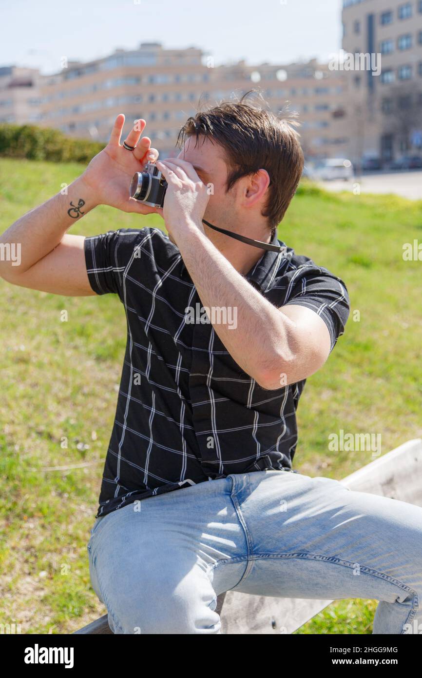 Junger Mann sitzt auf einer Bank und fotografiert im Garten mit einer Vintage-Kamera. Vertikales Shooting Stockfoto