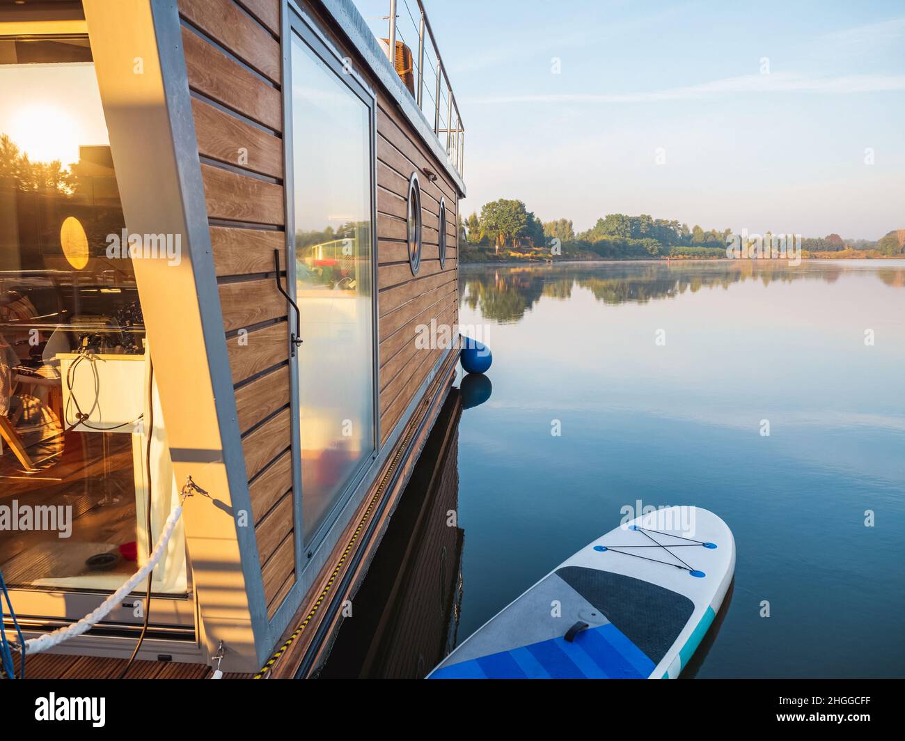 hausboot auf einem Fluss in einem frühen sonnigen Morgen auf einem Pier mit Paddleboard vertäut. Schwimmende Haus ist ein angenehmer Ort für das Wochenende zu mieten Stockfoto