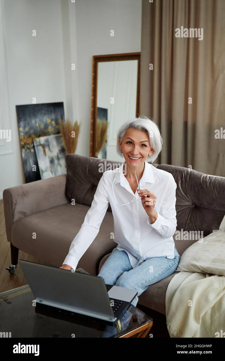 Positive Frau mit kurzen Haaren in formeller Kleidung, die auf einem weichen Sessel sitzt und die Kamera anschaut, während sie im hellen Raum einen Laptop durchstöbert Stockfoto