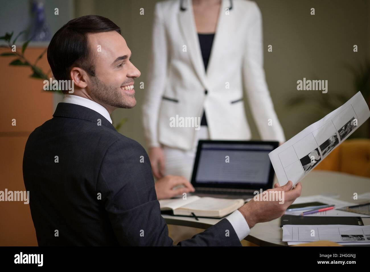 Geschäftsmann in einem klassischen Anzug und weißem Hemd sitzt in Profil zur Kamera an seinem Schreibtisch. Der Leiter des Unternehmens delegiert Aufgaben in seinem Büro. Arbeiter mit weißem Kragen.im Hintergrund ist eine Geschäftsfrau in einem weißen Anzug. Geröbter Hintergrund. Nahaufnahme. Hochwertige Fotos Stockfoto