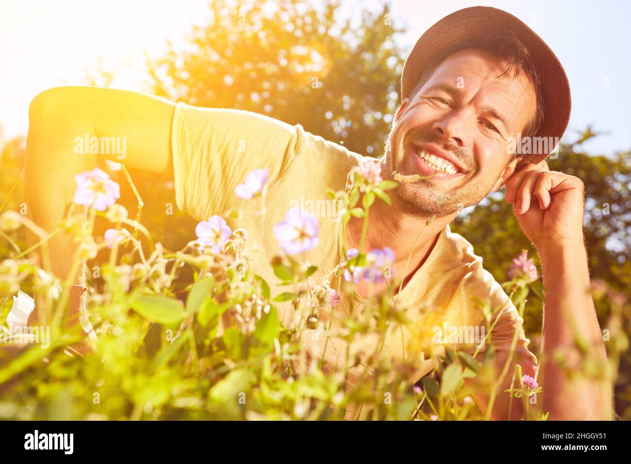 Glücklicher Mann im Hut, der im Sommer zwischen bunten Blumen auf der Wiese in der Natur sitzt Stockfoto