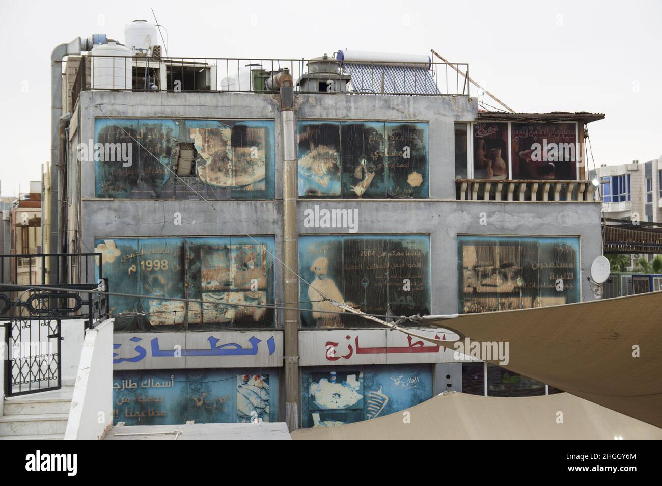 Verblasste und windgepeitschte Werbetafeln an einem Gebäude in Aqaba Jordanien, auf denen die Brotherstellung und ein Restaurant sowie arabische Schriftschnänge und zerrissene Markisen abgebildet sind. Stockfoto