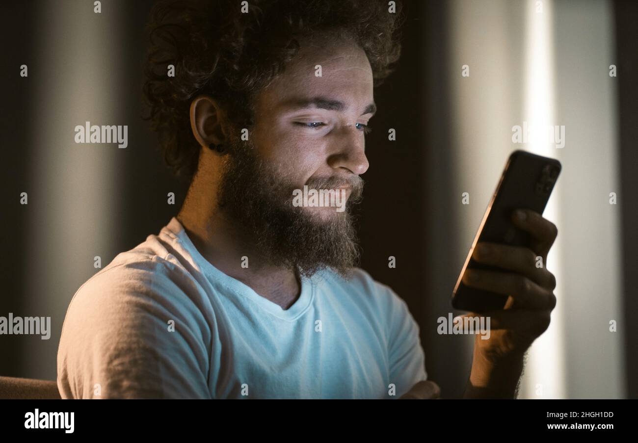 Curly Guy wipes auf seinem Telefon sitzen auf der Couch. Netter Mann tippt eine Nachricht an seine Freundin auf sein Telefon. Nahaufnahme im Hochformat. Hochwertige Fotos Stockfoto