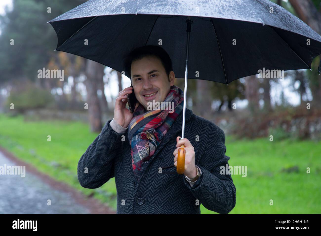 Junger eleganter Mann mit Regenschirm, der im Regen am Telefon telefoniert Stockfoto