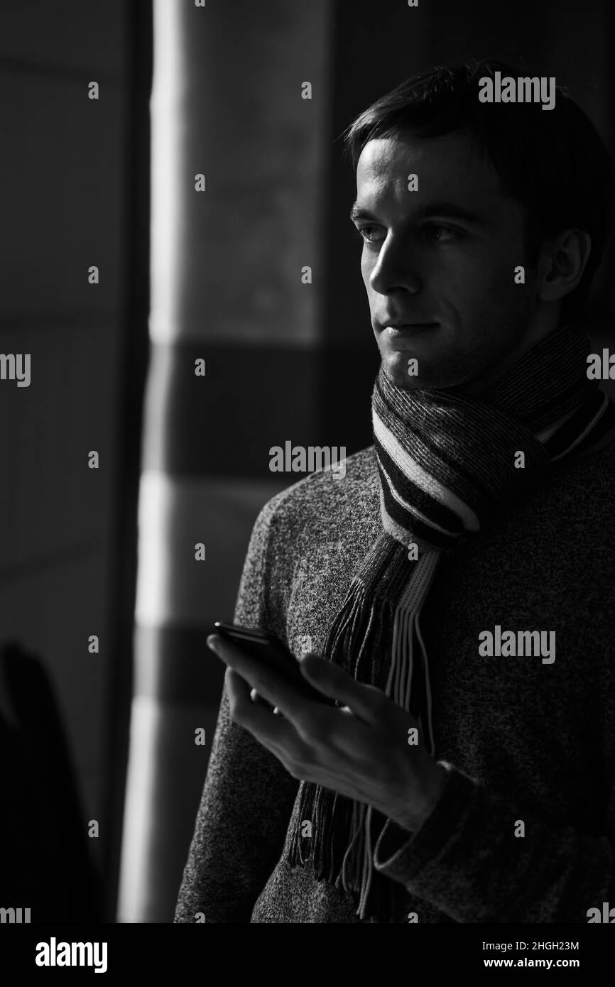 Ein junger, nachdenklicher, stilvoller Mann in einem modischen Pullover mit Schal steht neben dem Fenster und hält ein Telefon in den Händen und blickt auf den Bildschirm Stockfoto