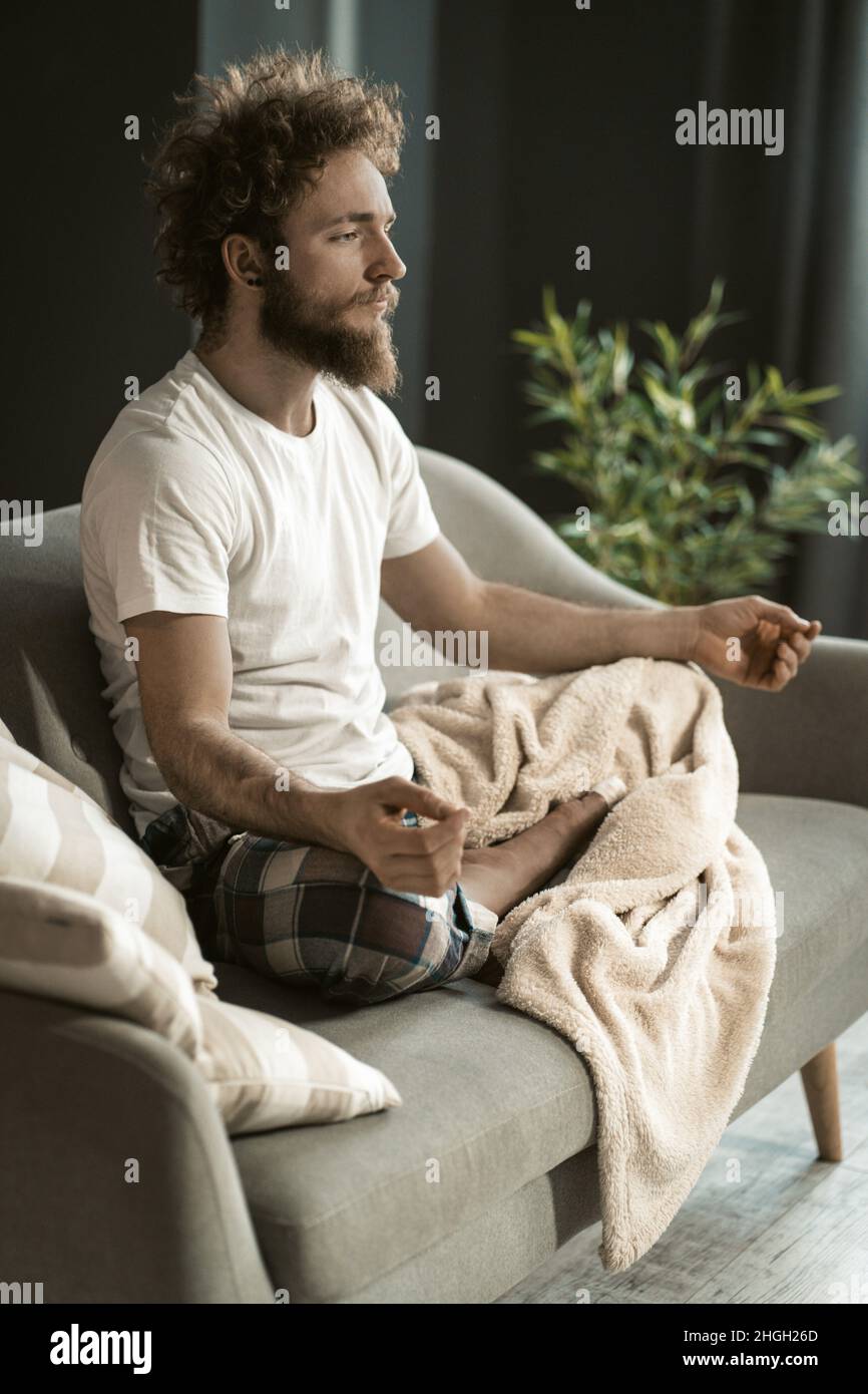 Junger entspannter Mann, der seinen täglichen Yoga auf dem Sofa in seiner Wohnung macht. Ein Mann mit betenden Händen und gefalteten Beinen, der in Lotus-Pose sitzt. Nahaufnahme. Hochwertige Fotos Stockfoto