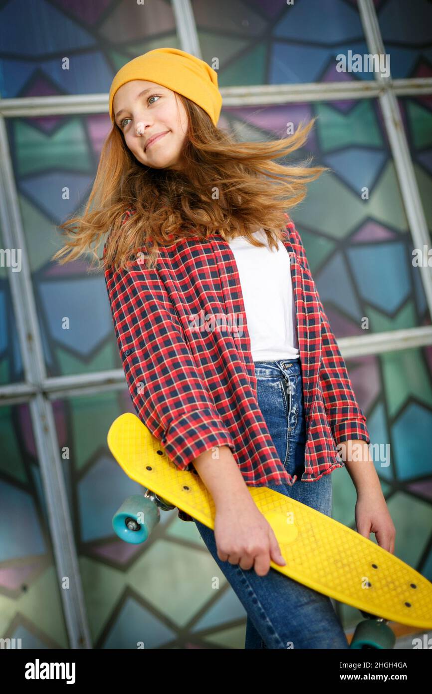 Lifestyle vertikale Outdoor-Porträt von jungen lächelnden Teenager-Mädchen mit einem gelben Skateboard tragen gelben Hut und roten karierten Hemd Stockfoto