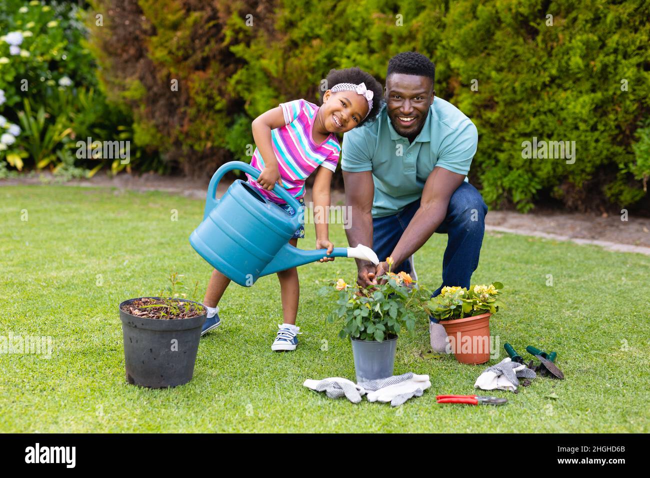Porträt eines lächelnden afroamerikanischen Mädchens, das Pflanzen vom Vater gießt, der im Garten kniet Stockfoto