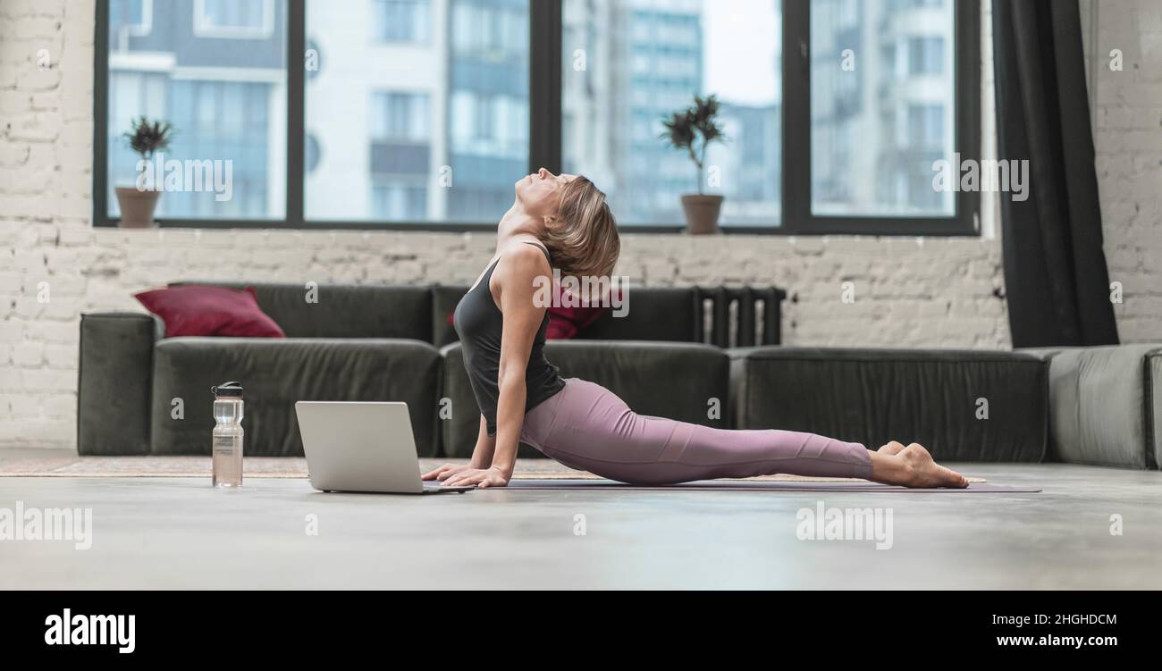 Die sportliche Frau sitzt auf einer Sportmatte und hat während der Quarantänezeit einen Online-Video-Chat mit ihrem Fitness Coach auf dem Laptop. Home Workouts. Stadthintergrund. Nahaufnahme. Hochwertige Fotos Stockfoto