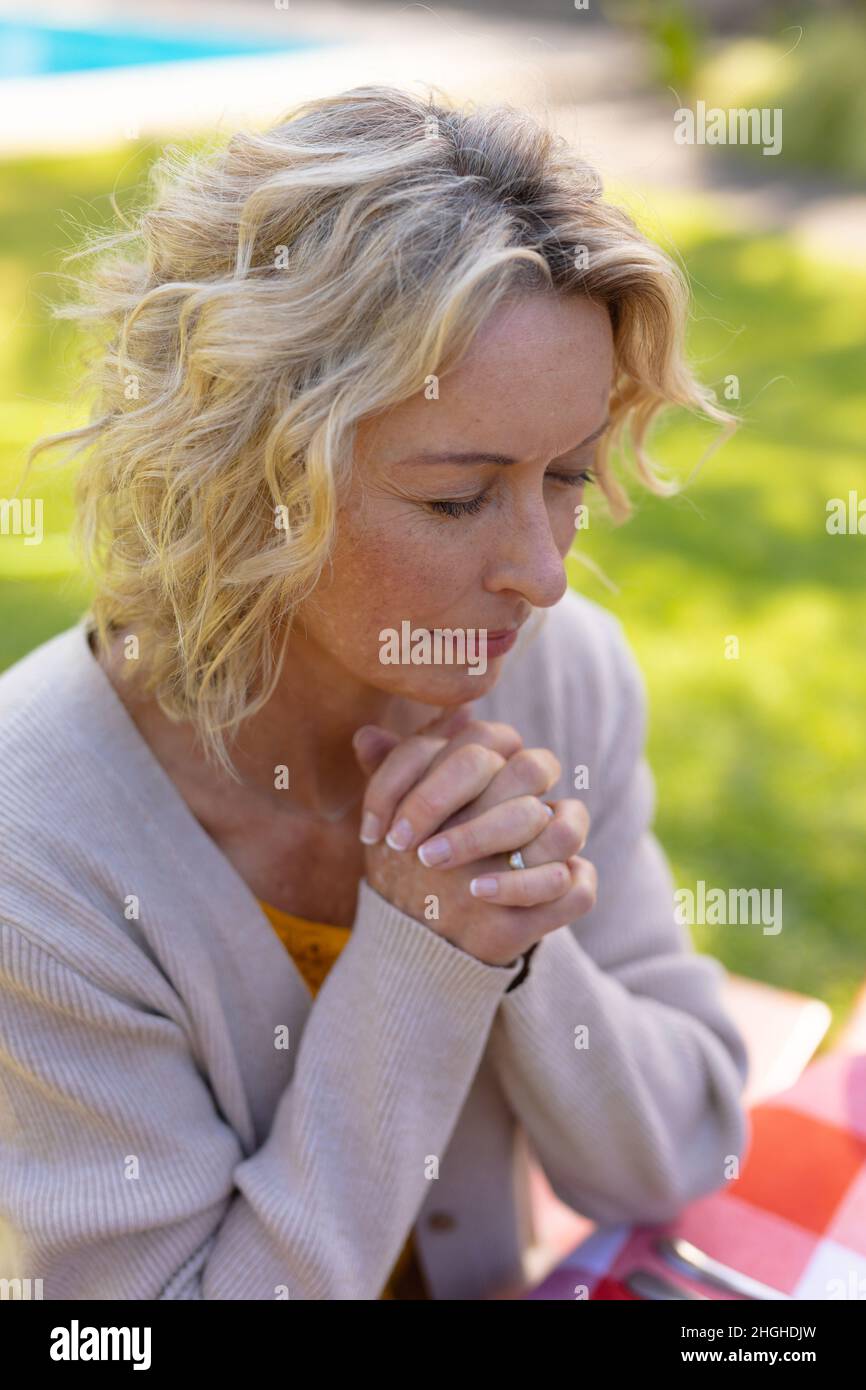 Blonde ältere kaukasische Frau mit geschlossenen Augen und geschlossenen Händen beim Beten im Garten Stockfoto