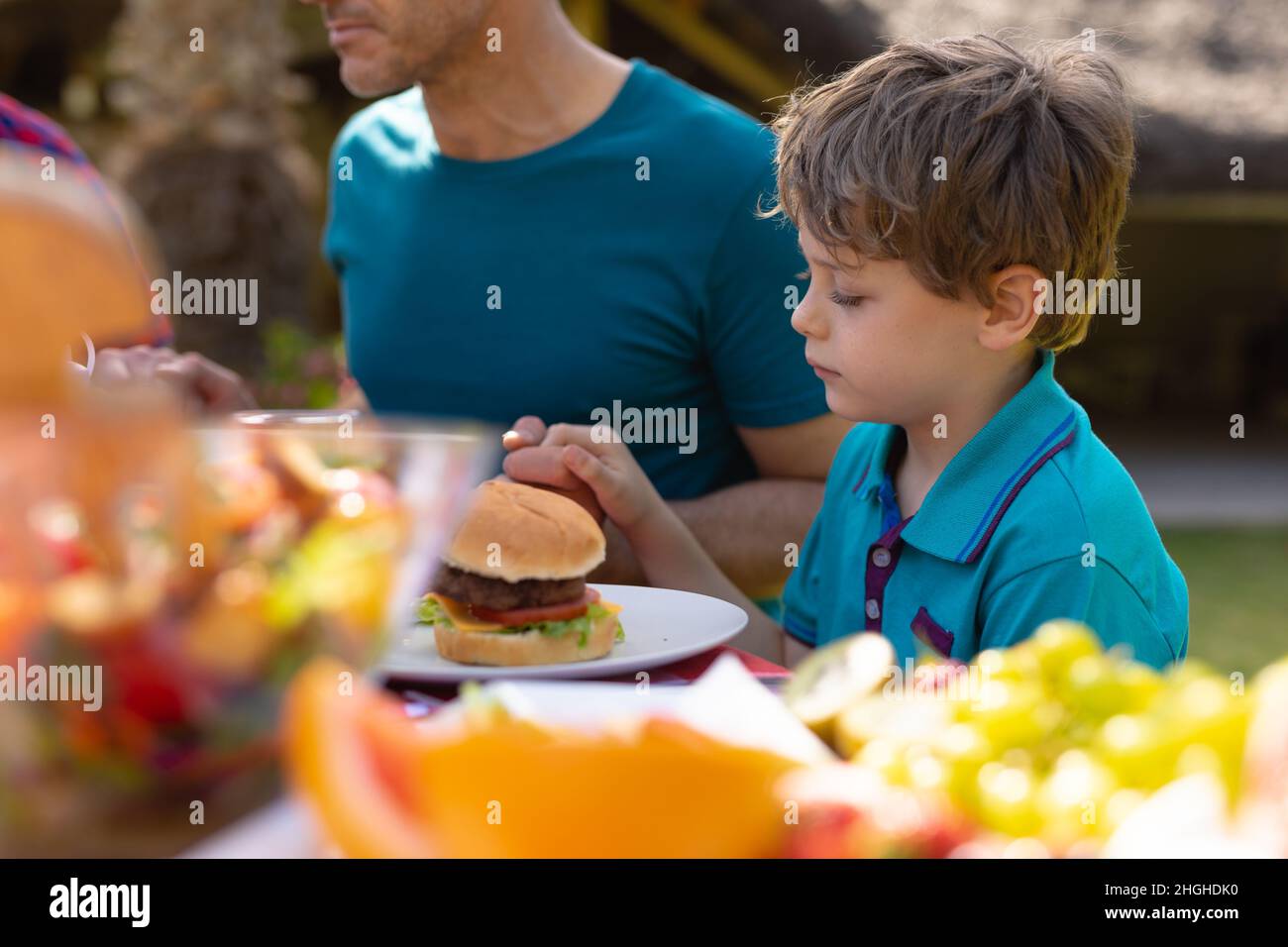 Kaukasischer Junge, der die Hand des Vaters beim Beten hält, während er mit Essen am Tisch im Garten sitzt Stockfoto