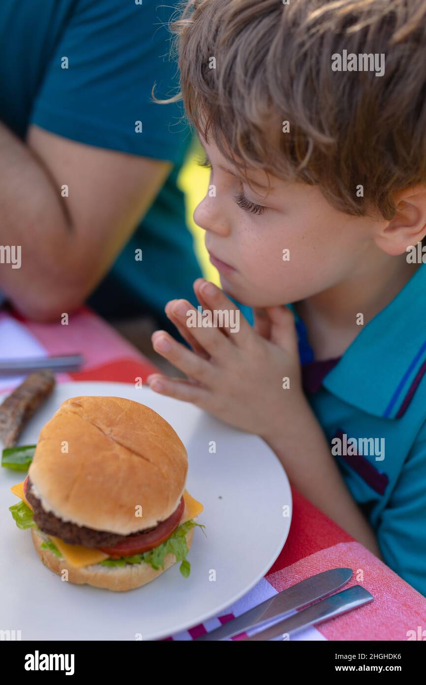 Kaukasischer Junge mit zusammengekrallten Händen und geschlossenen Augen und gebetet von einem Burger auf dem Tisch im Garten Stockfoto