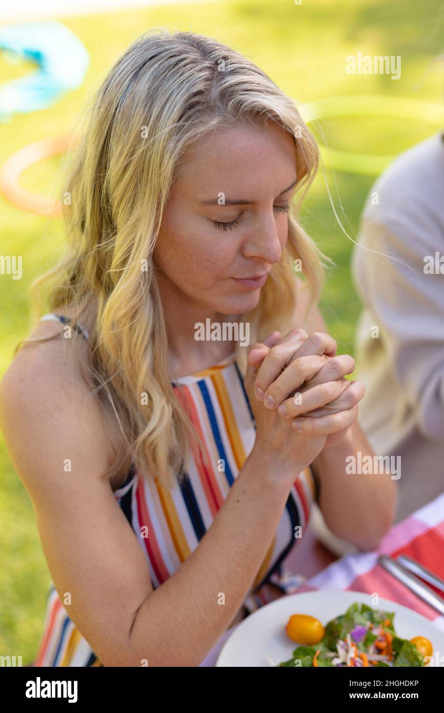 Blonde kaukasische Frau mit zusammengekrallten Händen und geschlossenen Augen beim Beten am Tisch Stockfoto