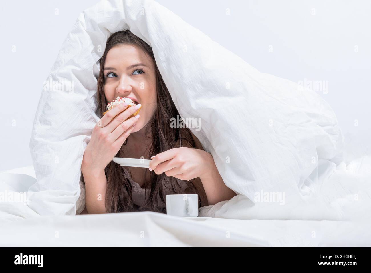 Das Mädchen isst heimlich Frühstück. Guten Morgen. Eine Frau liegt im Bett und beißt einen appetitlichen Cupcake ab, daneben gibt es Kaffee. Weißes Schlafzimmer. Stockfoto