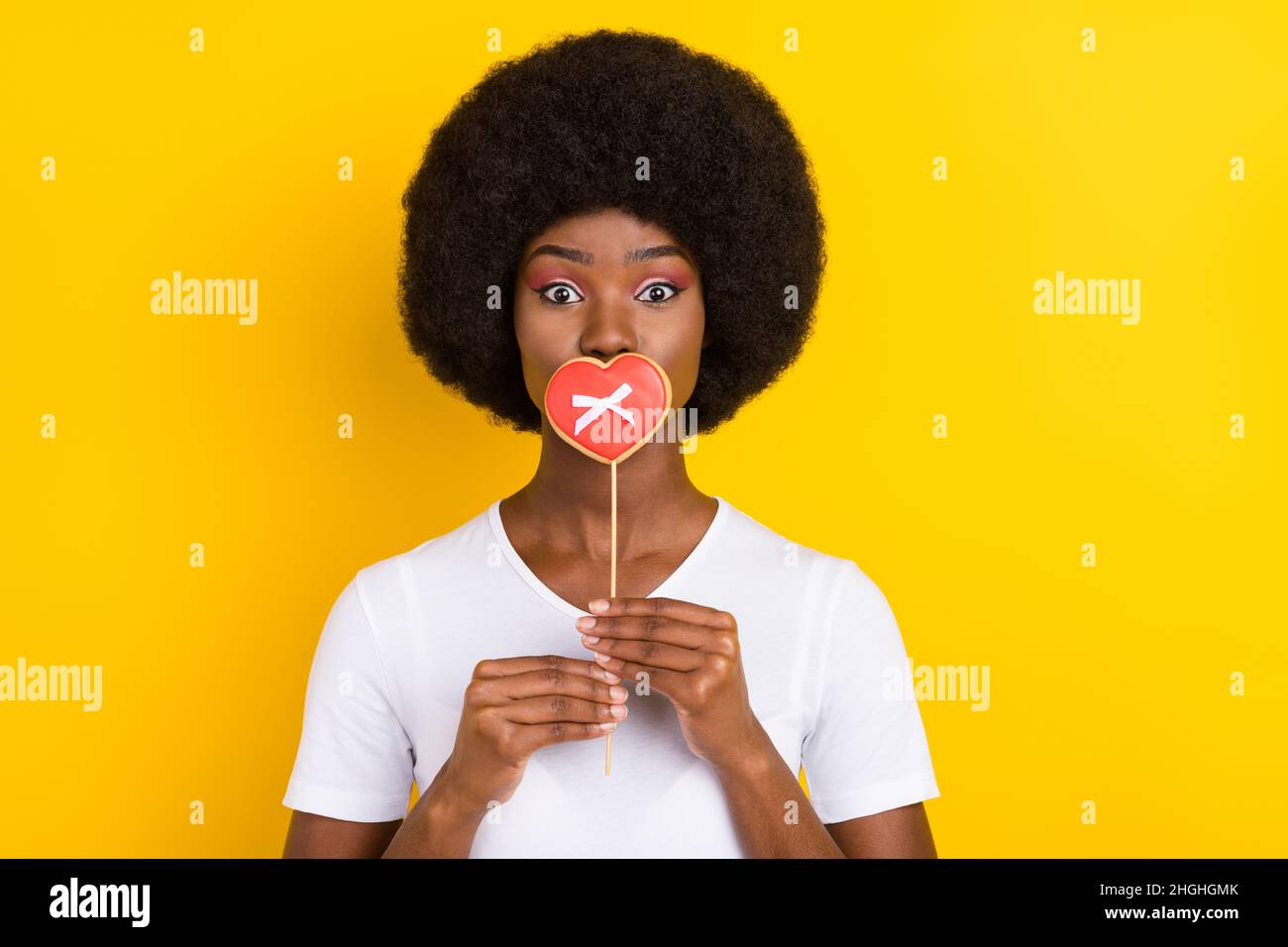 Foto von jungen funky afro amerikanische Frau decken Mund Süßigkeiten Herz Stick auf gelbem Hintergrund isoliert Stockfoto