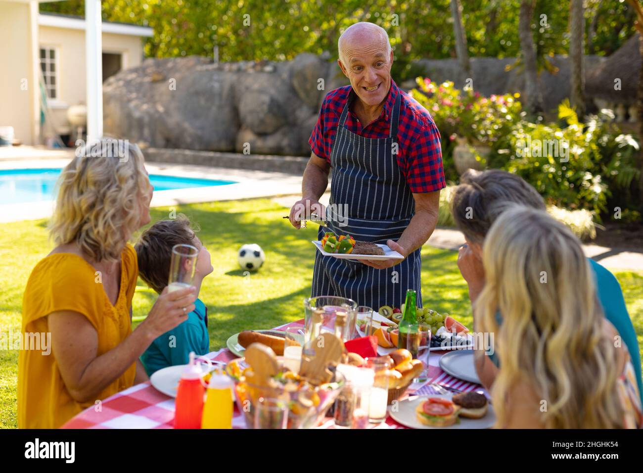 Glücklicher kaukasischer älterer Mann, der der Familie Grillgerichte serviert, die zusammen am Tisch im Garten sitzt Stockfoto