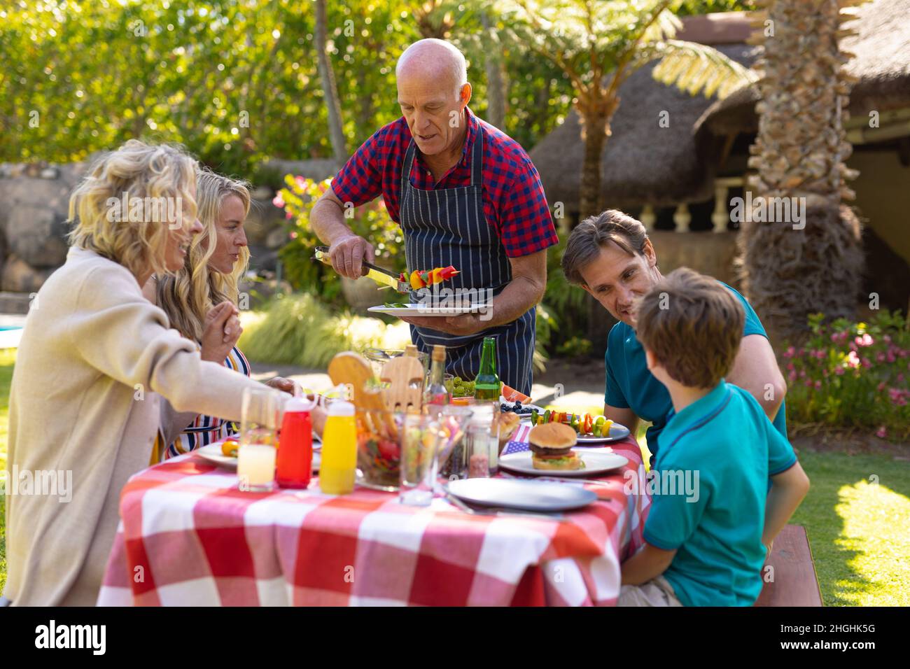 Kaukasischer älterer Mann, der der Familie Grillgerichte serviert, die zusammen am Tisch im Garten sitzt Stockfoto