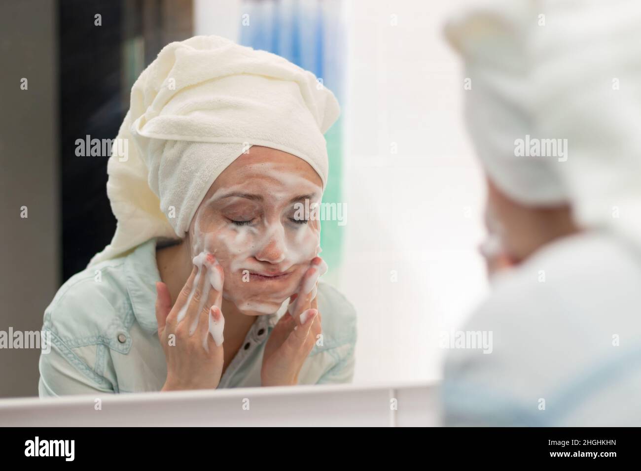Eine junge Frau mit einem weißen Handtuch auf ihrem Kopf im Badezimmer wäscht eine Gesichtsmaske ab. Spiegelung im Spiegel. Selektiver Fokus. Nahaufnahme. Hochformat Stockfoto