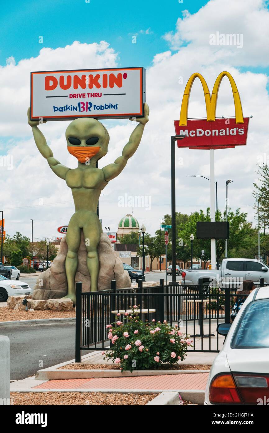 Eine Alien-Statue hält ein Dunkin' Donuts-Schild in Roswell, New Mexico, während sie eine Gesichtsbedeckung trägt. McDonalds UFO Restaurant ist sichtbar hinter Stockfoto