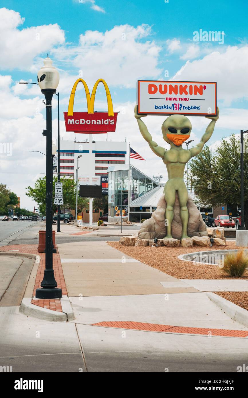 Eine Alien-Statue hält ein Dunkin' Donuts-Schild in Roswell, New Mexico, während sie eine Gesichtsbedeckung trägt. McDonalds UFO Restaurant ist sichtbar hinter Stockfoto