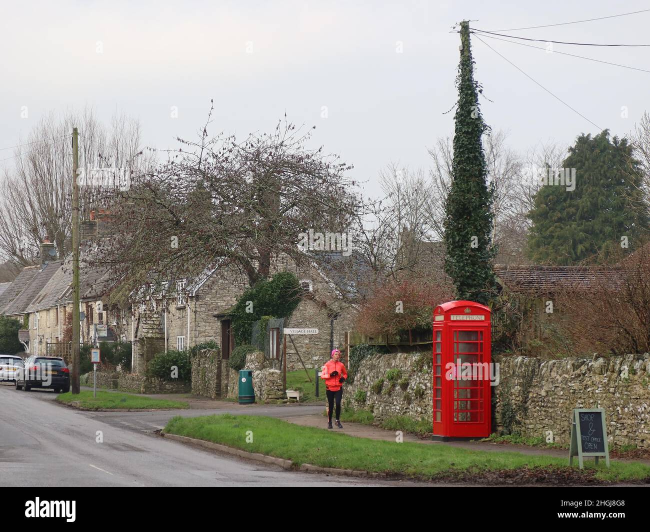 Eine Joggerin in rotem Mantel, rotem Hut und schwarzen Handschuhen kommt an einer roten Telefonbox im Dorf Martinstown Dorchester Dorset vorbei Stockfoto